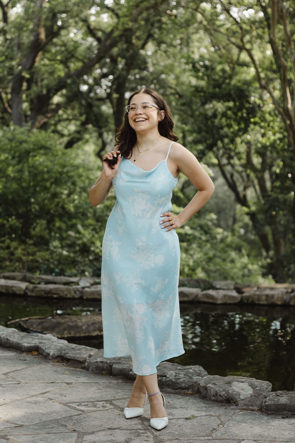 A light blue floral dress is showcased in a natural garden setting with stone path near a tranquil pond.