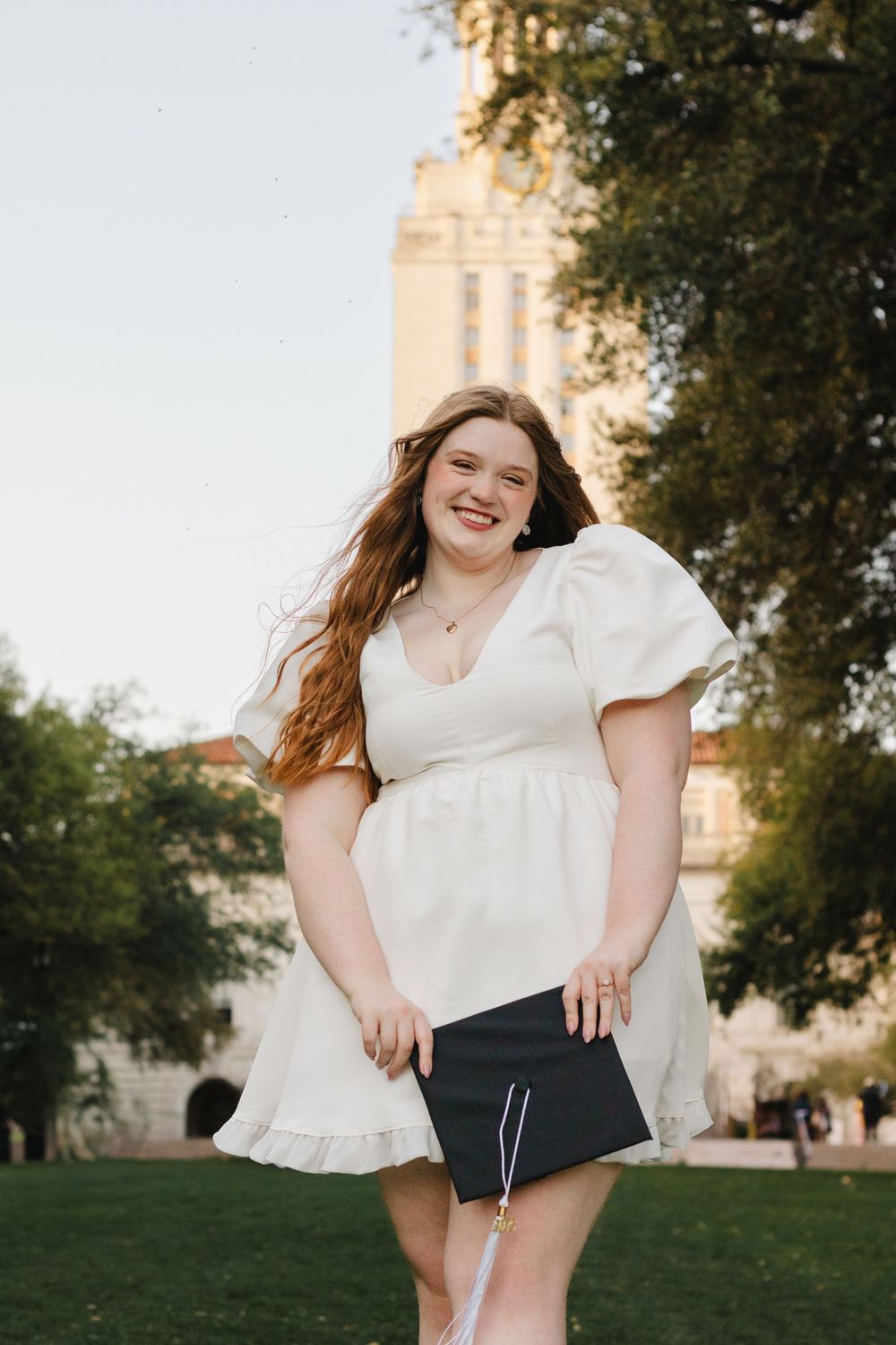 A graduate wearing a white dress holds a diploma while standing on campus grounds at sunset.