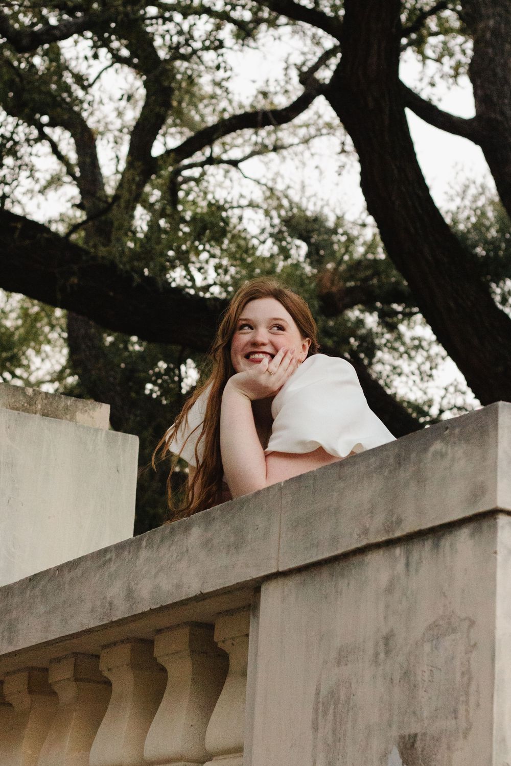 Woman in white leaning on concrete balcony railing with trees in background during outdoor portrait session.
