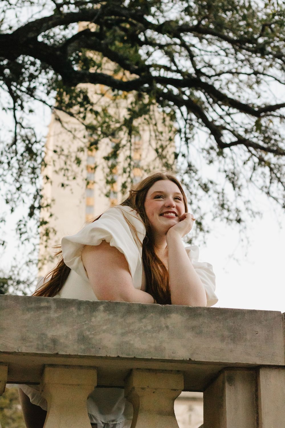 A person in a white top leans over a stone balcony, smiling while surrounded by overhanging tree branches.