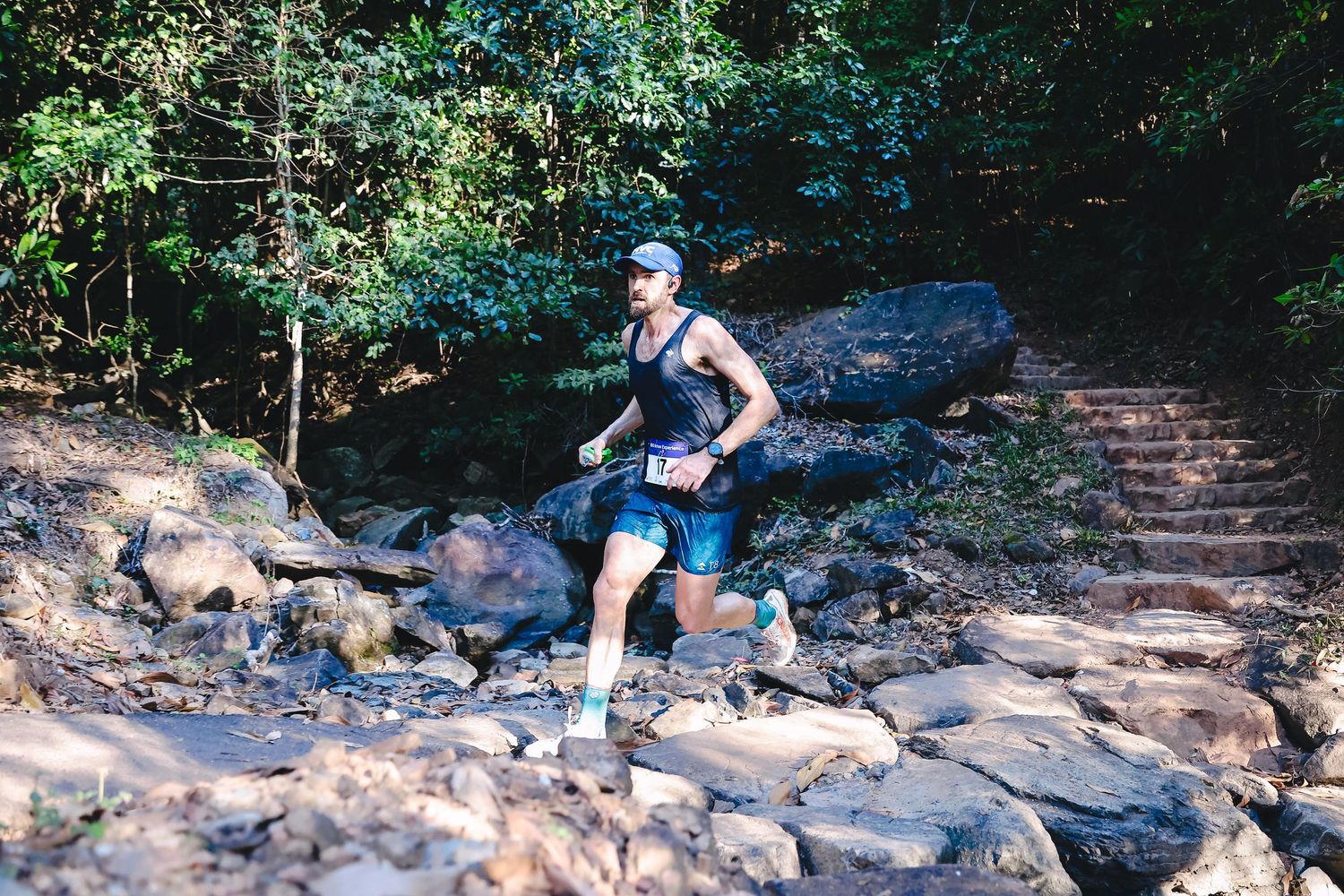 A trail runner navigates along a rocky dirt path through dense forest vegetation on a nature trail.