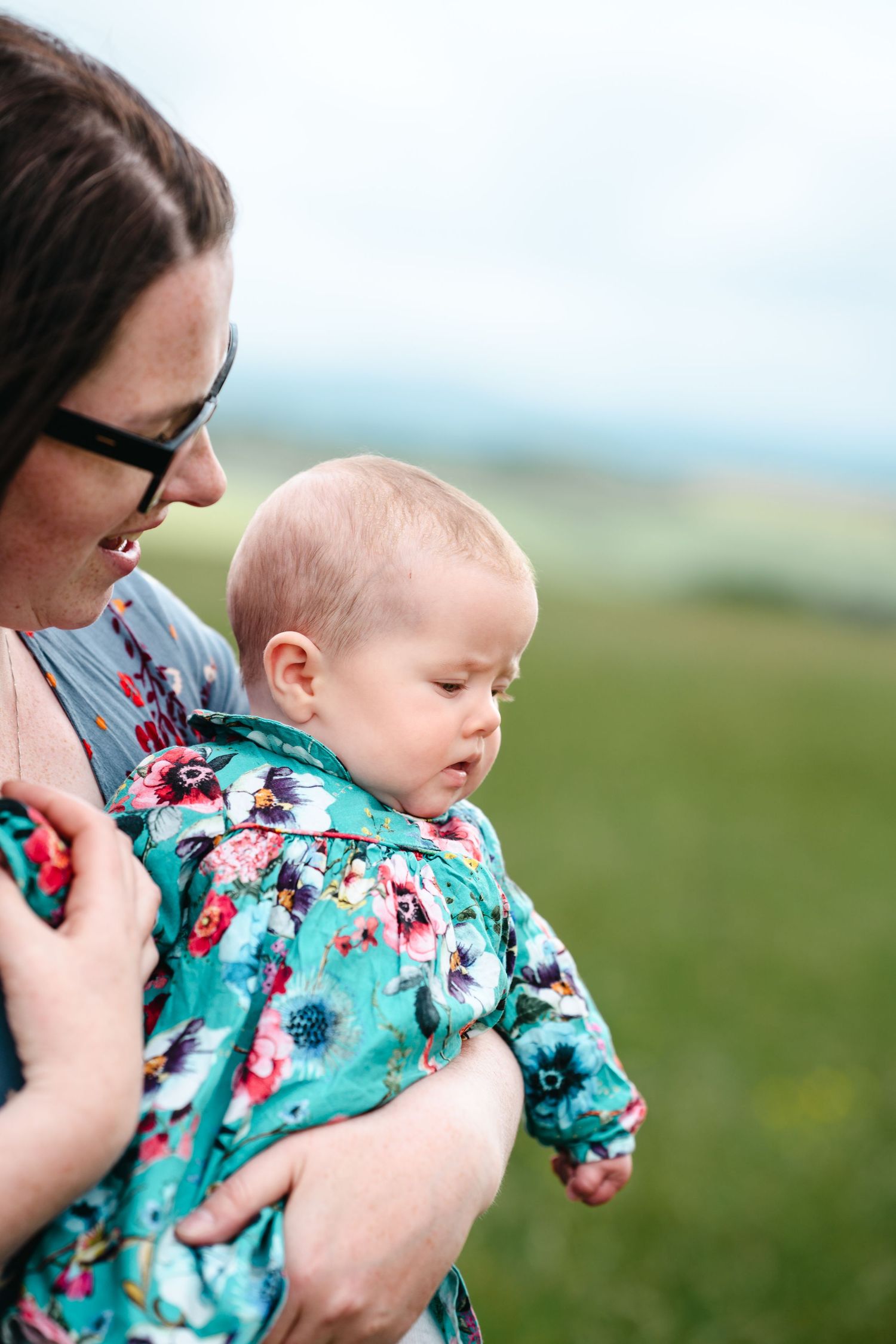 Mother holds baby wearing floral outfit against green outdoor background.