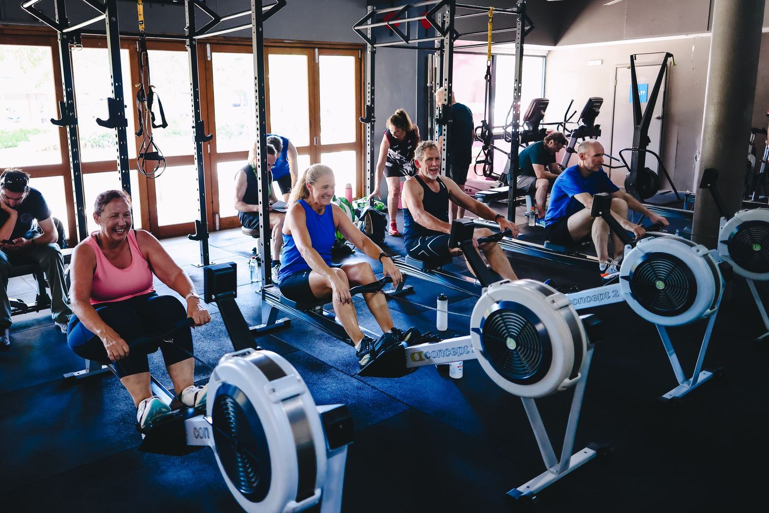 People exercising on rowing machines at a gym with fitness equipment and mirrors in the background.