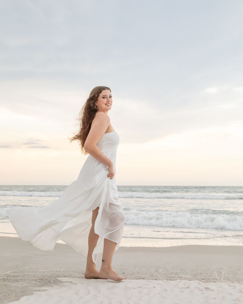 Woman in flowing white dress twirls on sandy beach at sunset as ocean waves roll in the background.