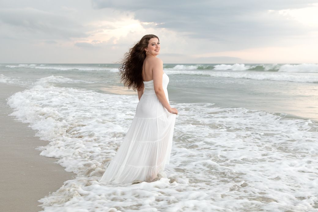 A bride in a white flowing dress stands at the edge of ocean waves on a sandy beach at sunset.