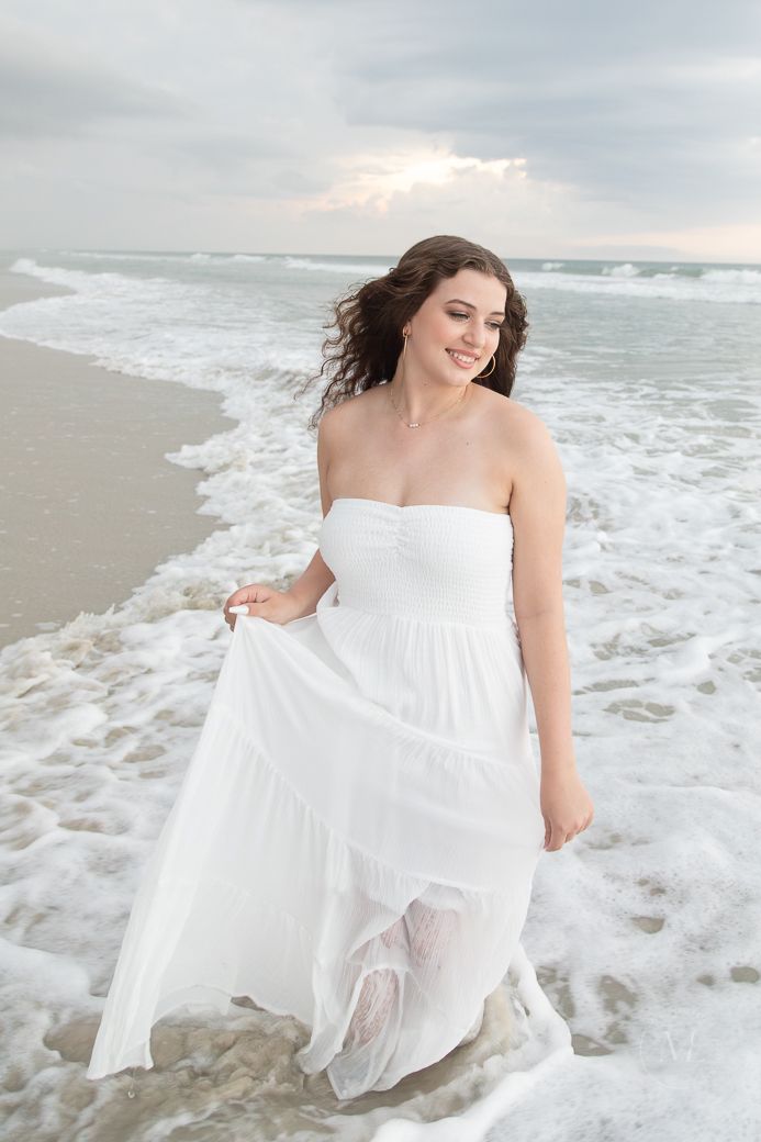 Flowing white dress billowing in ocean waves during a beachside photoshoot at sunset.