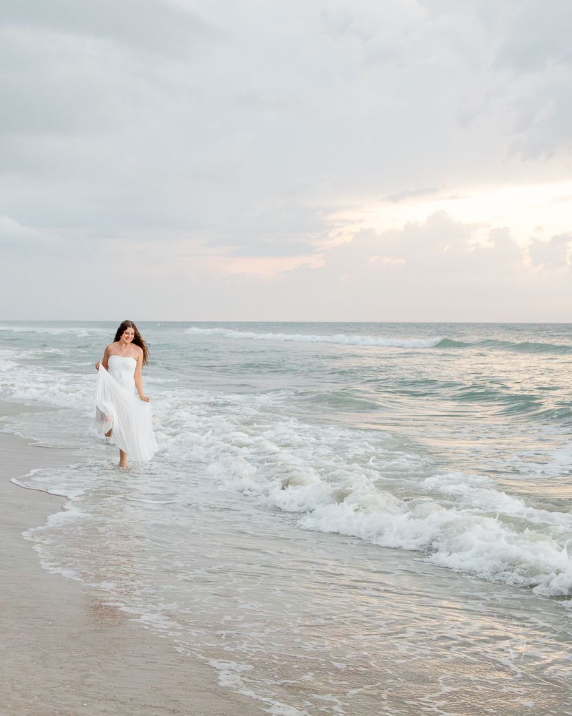 A flowing white dress contrasts against crashing ocean waves during a serene beach photoshoot at sunset.