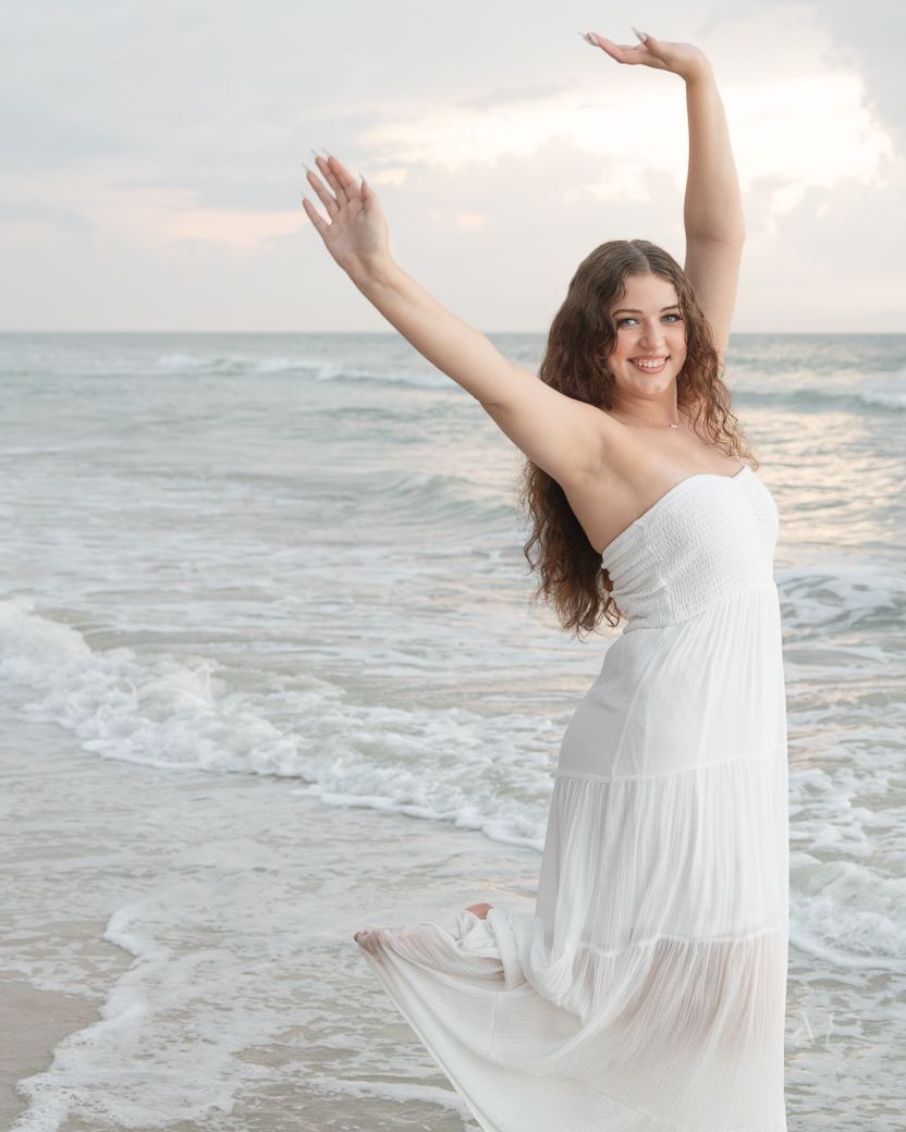 A carefree moment captured on a beach with waves and clouds as someone in white raises their arms joyfully.