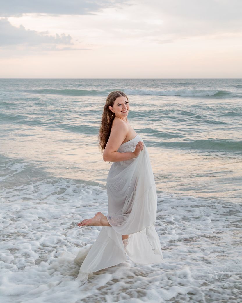 A flowing white dress catches the ocean breeze during a sunset photoshoot on the beach.
