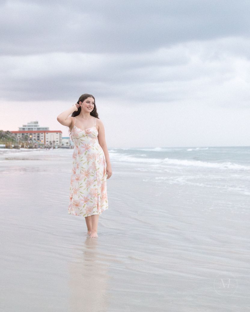 A floral dress flows in the sea breeze as a beachgoer stands in shallow ocean water at sunset.