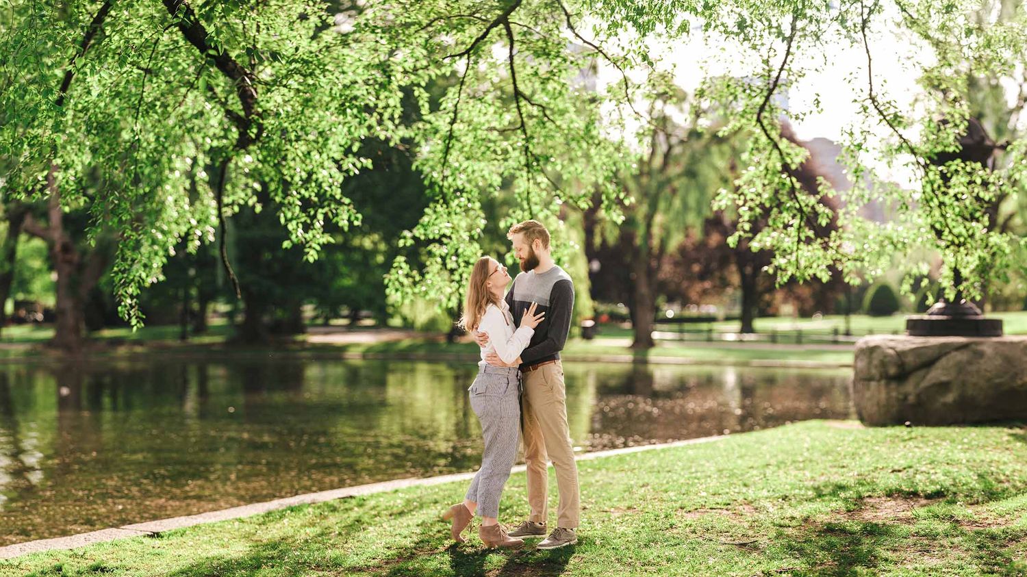 Wide angle portrait of engaged couple standing by Lagoon under sweeping branches in iconic Boston Public Garden engagement session