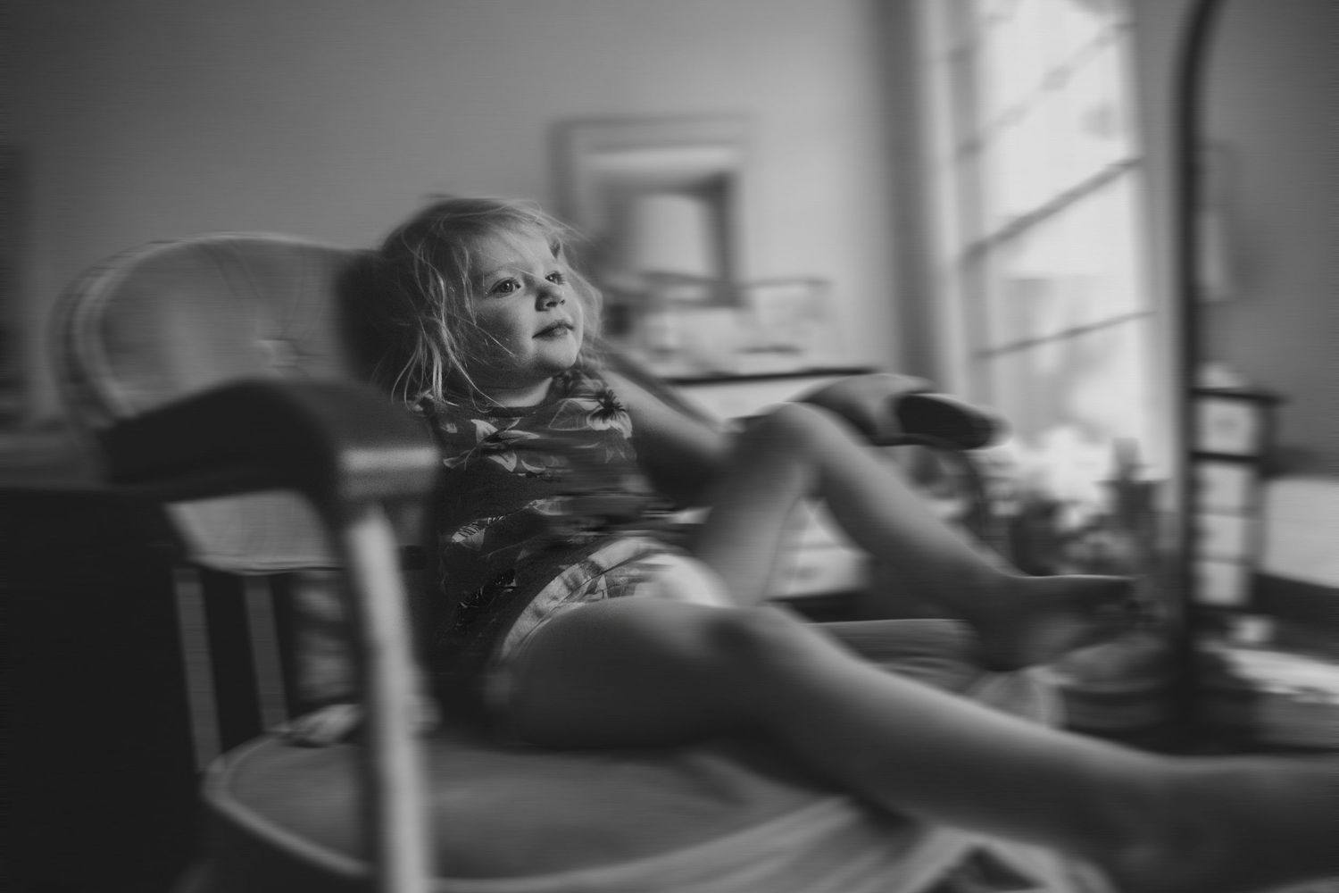 Young child relaxes in spinning office chair by window in black and white candid photograph.