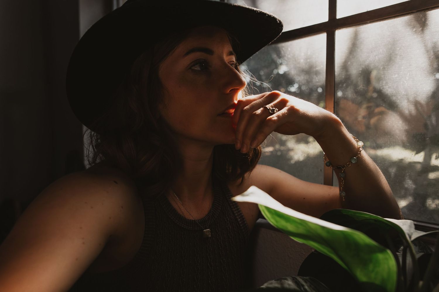 A silhouette of a woman by the window with her hand to her mouth while soft light streams through the glass.