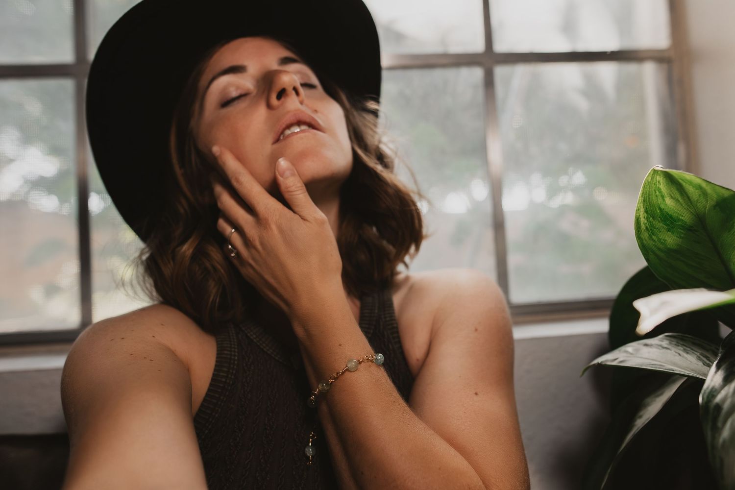 Person in a black hat poses near a window with natural light and indoor plants, creating a moody aesthetic.