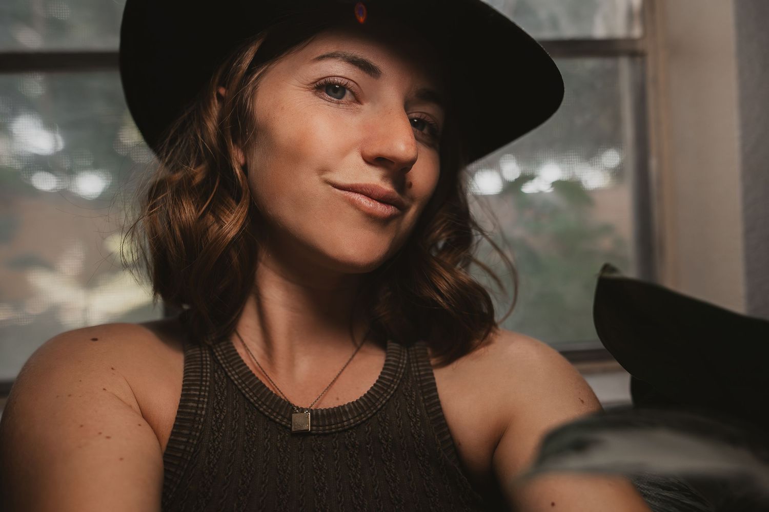 Woman poses seductively in black hat near window with moody lighting in vintage inspired portrait series.