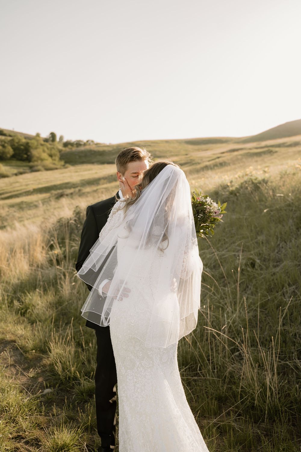 A couple shares an intimate moment in their wedding attire on a golden hillside at sunset.