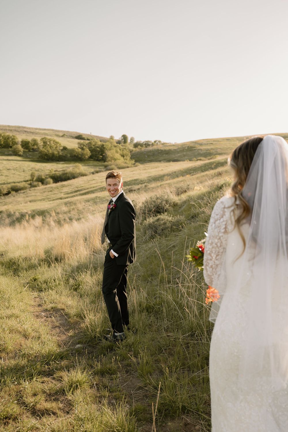 A wedding couple stands in tall grass on a hillside during golden hour.
