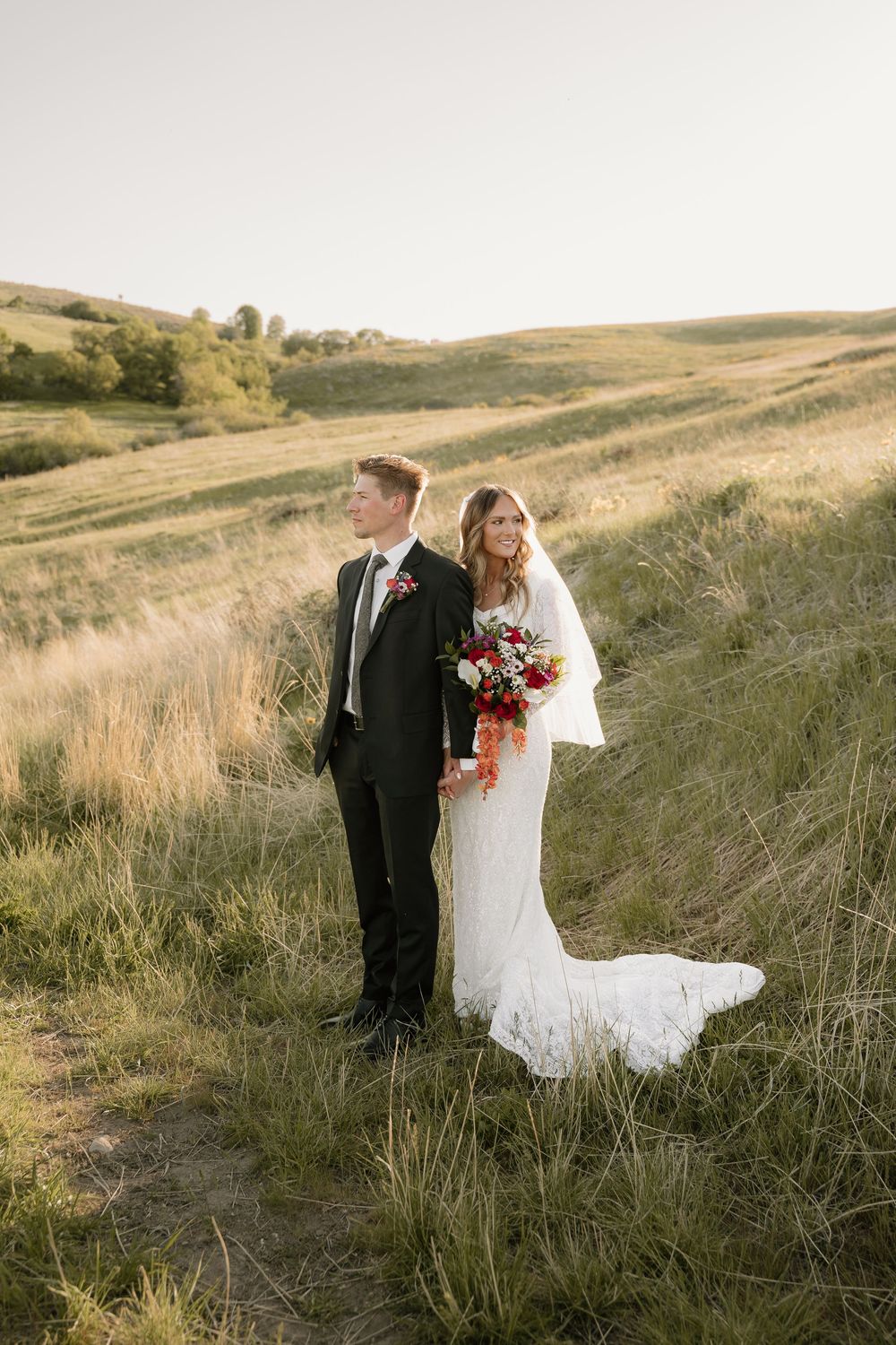A newlywed couple poses in a wedding dress and suit on a scenic hillside during golden hour.