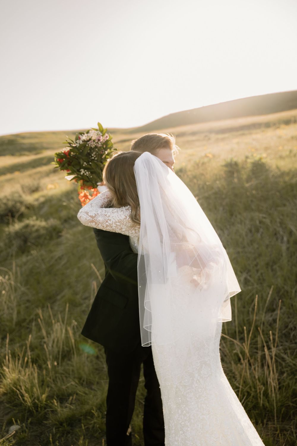 A bride and groom embrace in a romantic sunset moment on a grassy hillside with soft golden light.