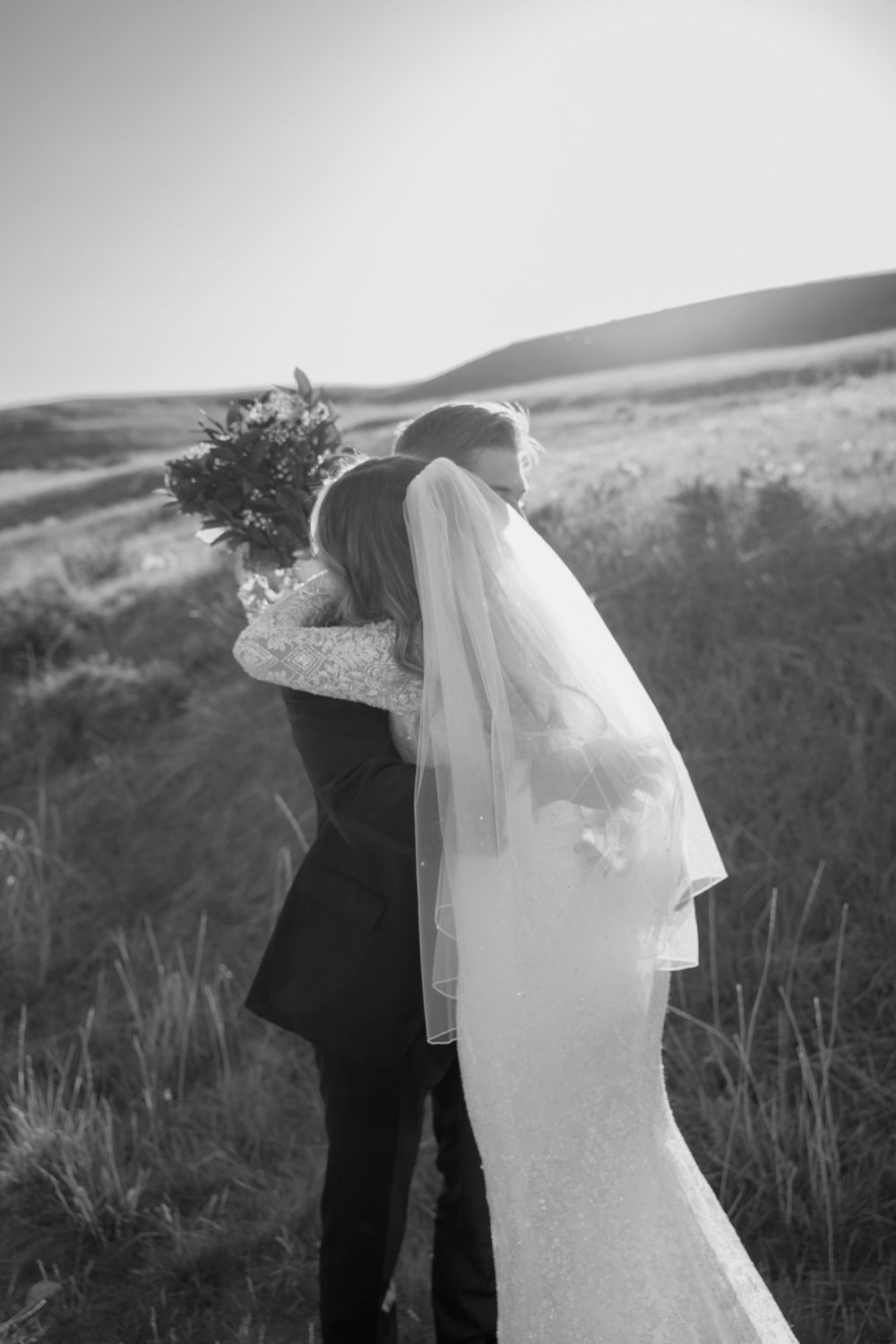 A romantic black and white wedding photo of a couple embracing in a scenic hillside meadow.