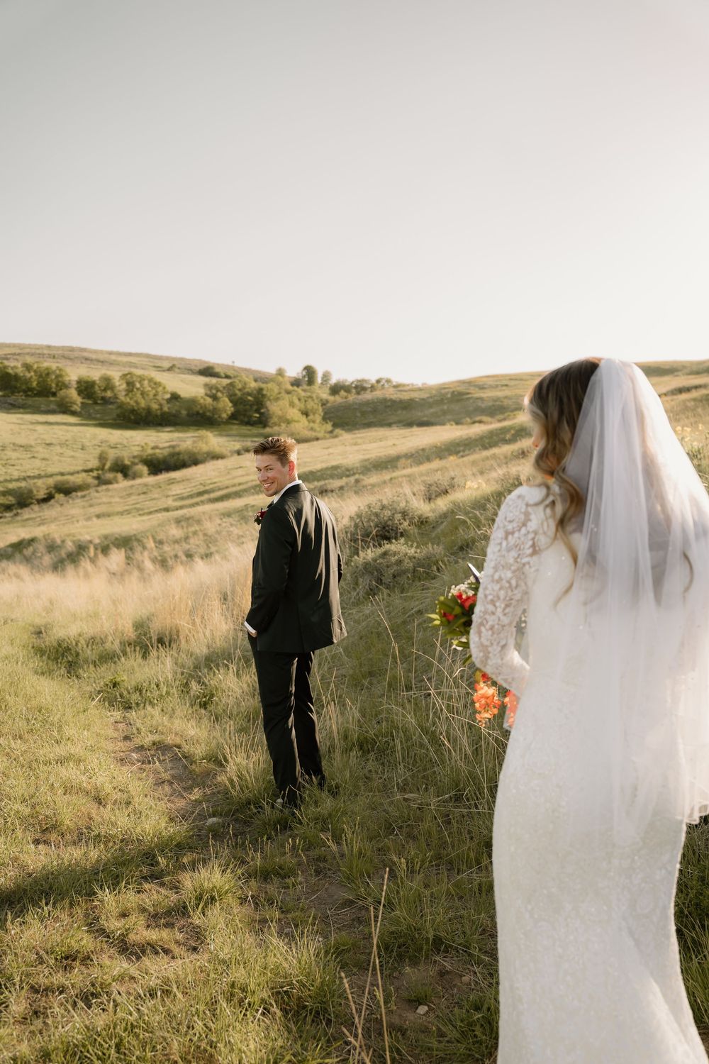 A bride in a lace dress and veil on a grassy hillside during a golden hour wedding photoshoot.