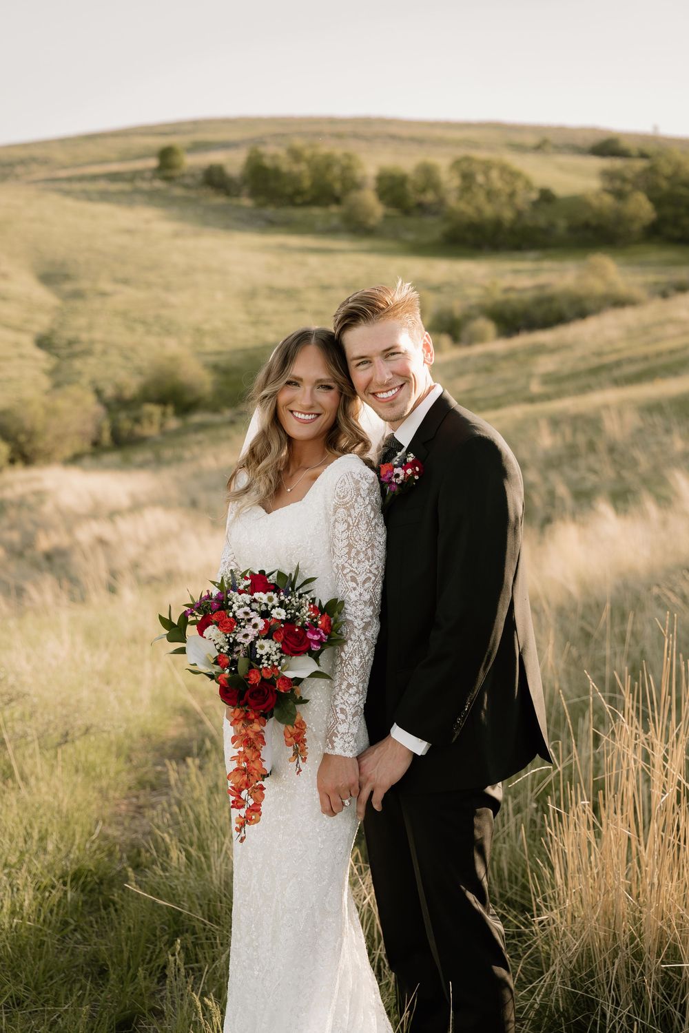 A happy couple in wedding attire pose together against a golden hillside landscape during sunset.