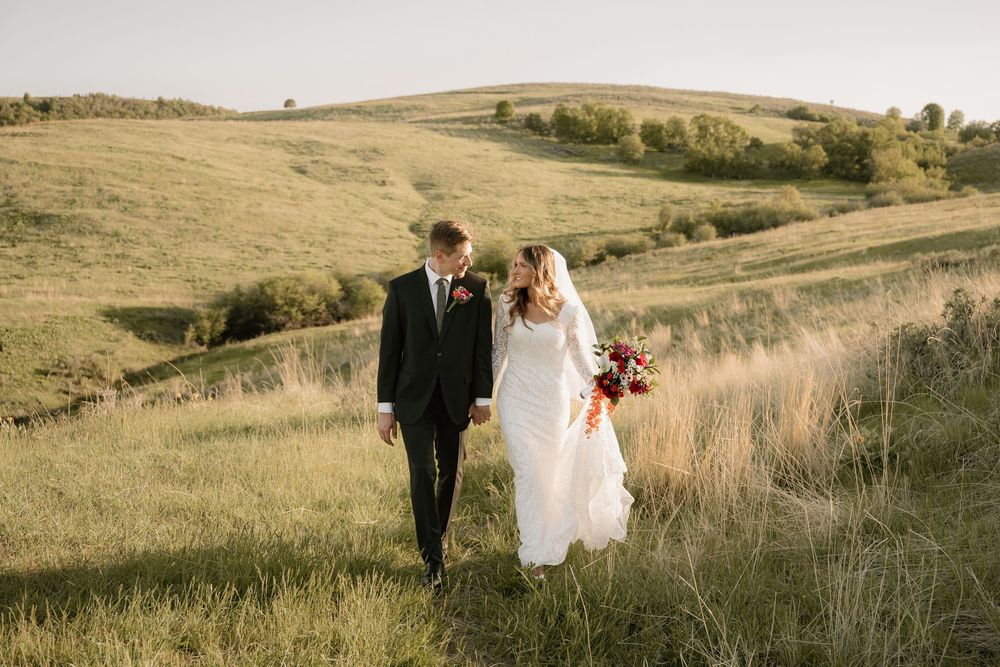 A couple walking together through tall grass on their wedding day with rolling hills in the background.