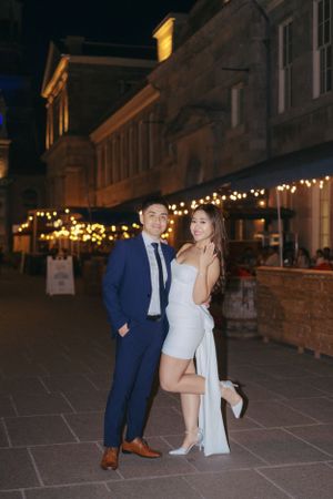 Couple posing under string lights on historic street at night.