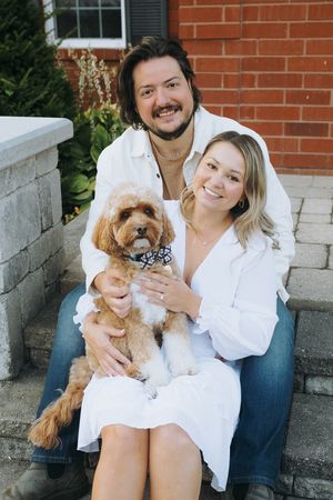 A couple poses on steps with their adorable goldendoodle puppy wearing a bandana.