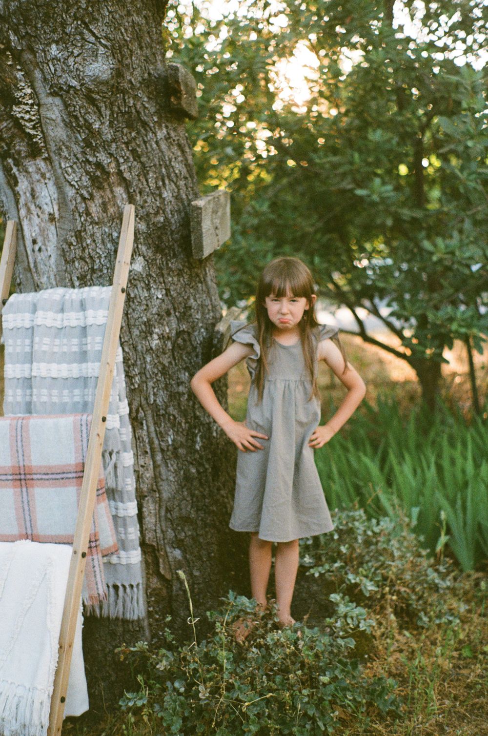 A child poses confidently in a gray dress next to a large tree with blankets hanging nearby.