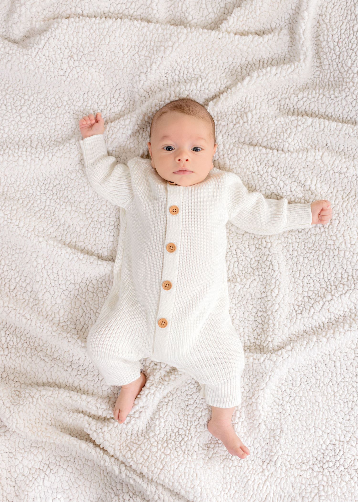 Baby wearing white knitted romper with wood buttons lies on textured white blanket.