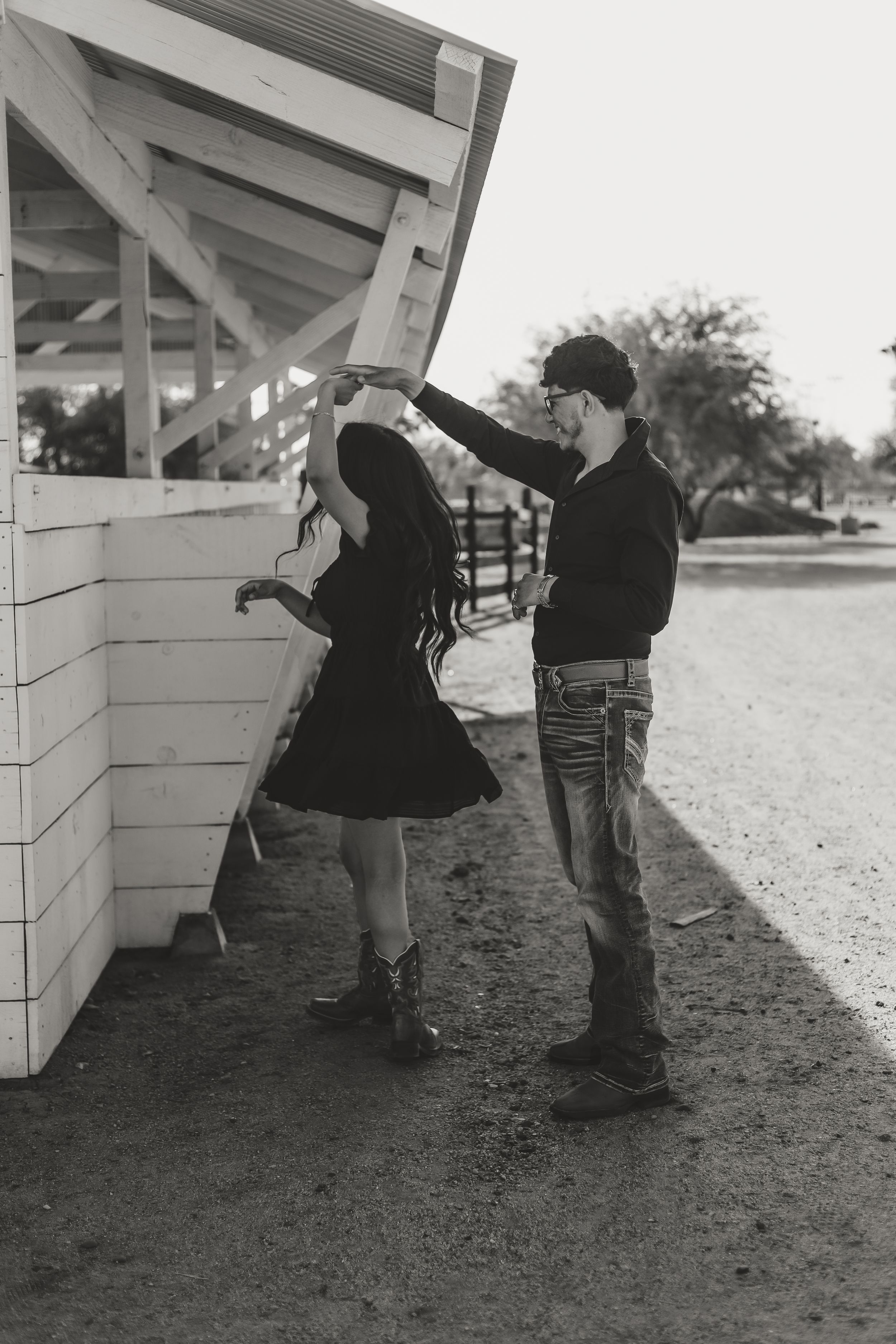 A black and white photo of two people in cowboy boots dancing together along the exterior of a barn structure.