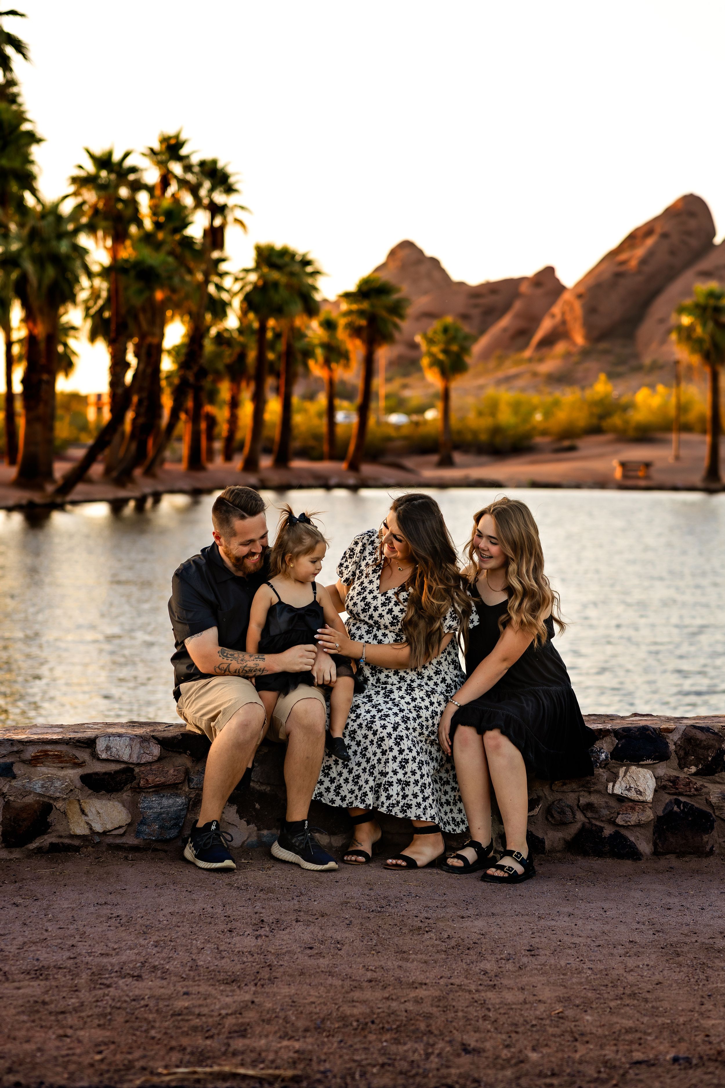 A family enjoys a sunset moment by a lakeside with palm trees and red rock formations in the background.