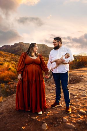 A couple stands together on a rocky mountain trail at sunset, with rich autumn colors surrounding them in the landscape.