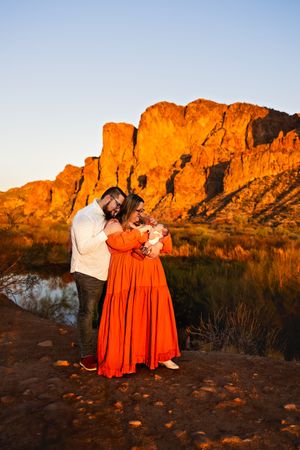 A couple embraces at sunset against towering red rock cliffs in the desert, with the woman wearing a vibrant orange dress.