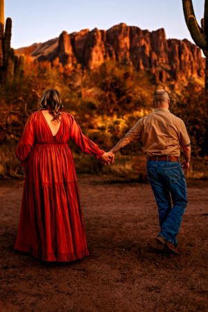 A couple in jeans and red dress walk away towards desert mountains at sunset with dramatic shadows in Arizona.