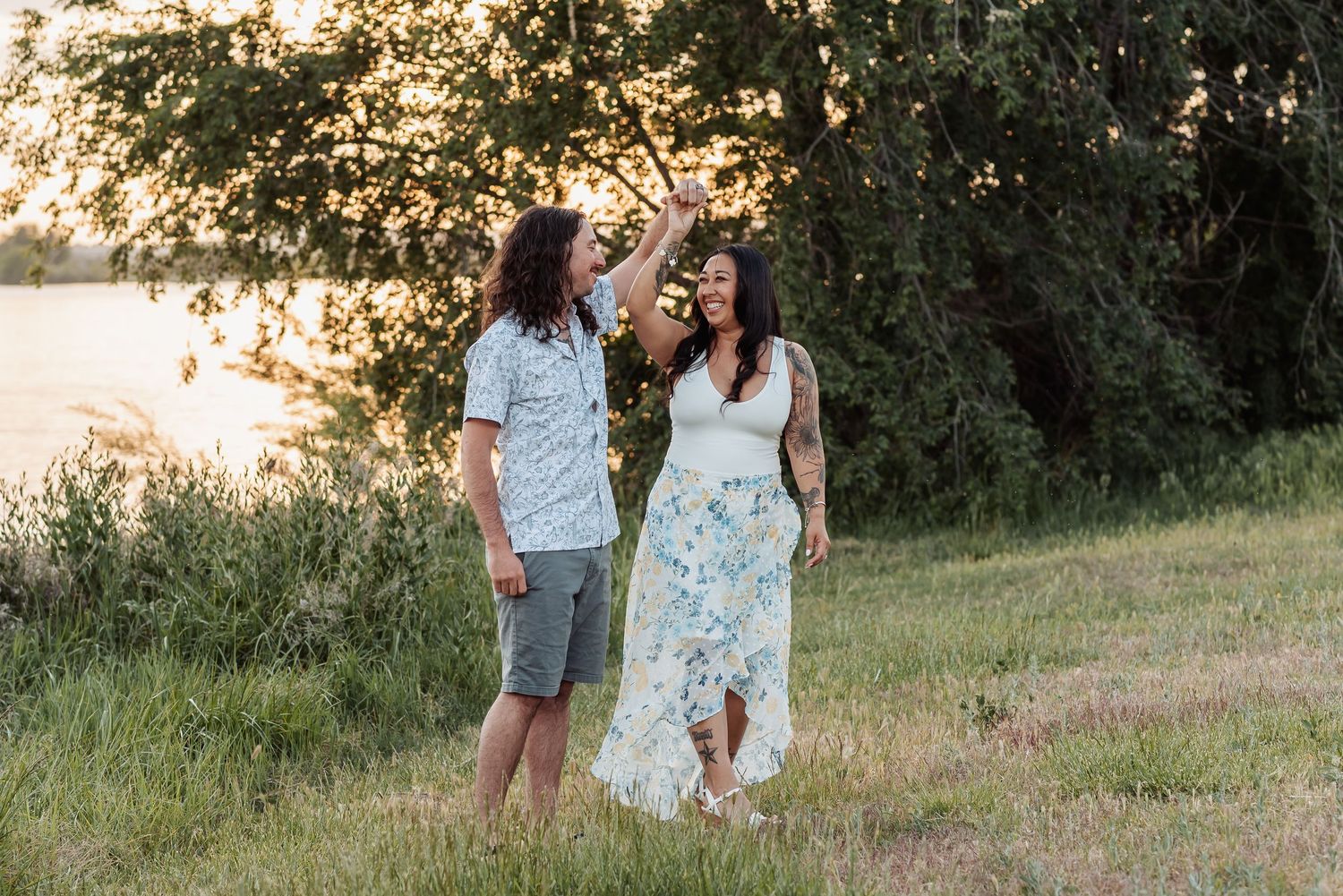 A couple stands together in a sunlit meadow during golden hour with green trees in the background at Chiawana Park in Pasco, Washington.