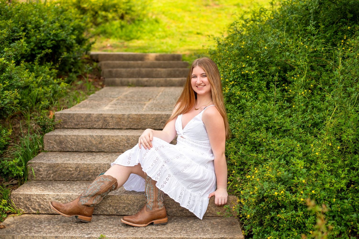 Someone in a white sundress and boots sits on rustic outdoor stone steps surrounded by lush green foliage.