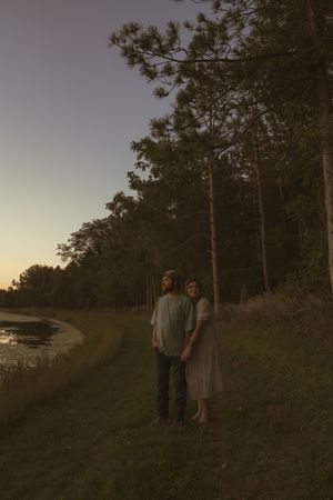 Two people standing on a hillside trail with pine trees and water views at dusk.