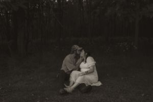 A black and white photo captures a tender moment between a couple sitting in a grassy field.