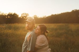 A couple embraces in warm evening light while standing in a tall grass meadow.