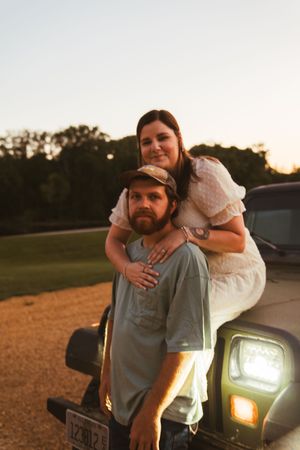 A couple poses playfully near their vehicle at sunset.