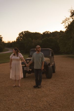 A couple stands hand in hand in front of their vehicle during sunset.