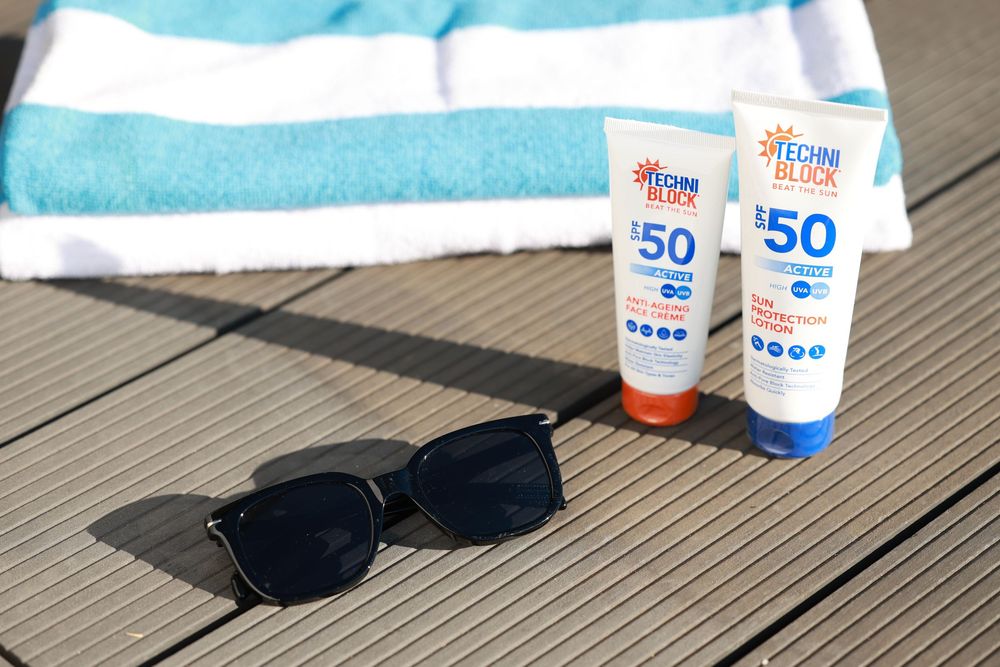 Two SPF 50 Techni Block sunscreen tubes beside black sunglasses on a wooden deck with a striped beach towel.