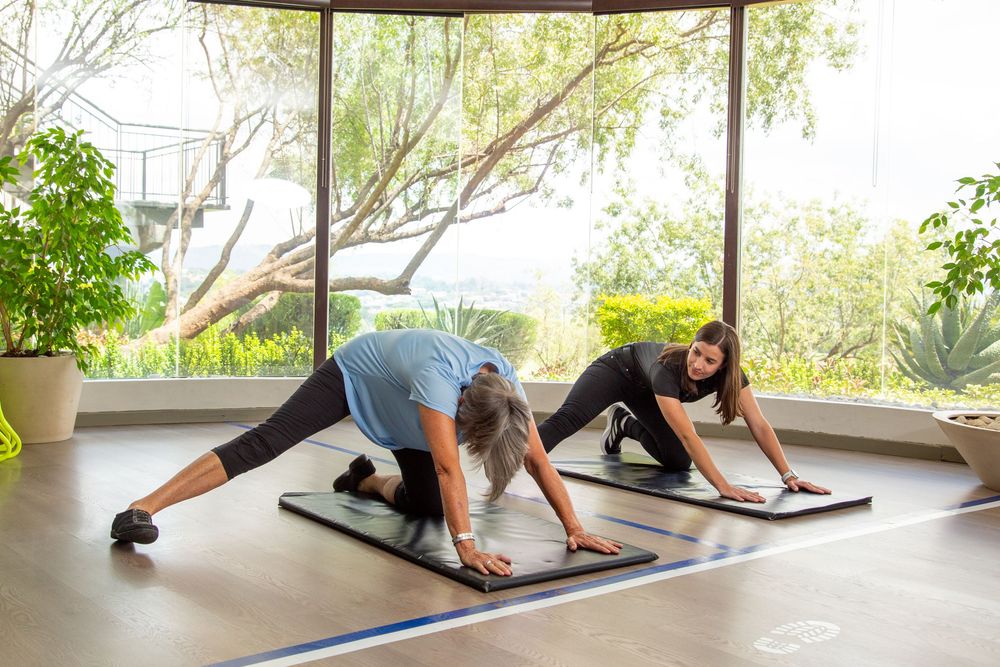 Two people practicing yoga stretches on exercise mats in an open-air fitness studio with natural lighting.