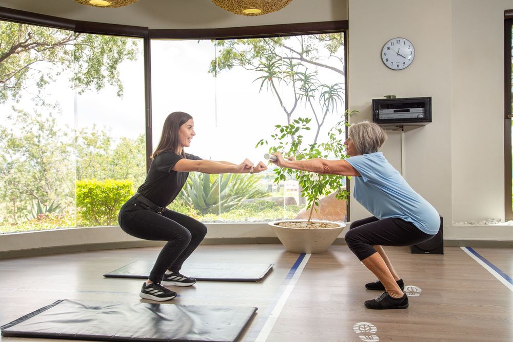 Two people perform synchronized squats together during a workout session in a bright fitness studio with large windows.