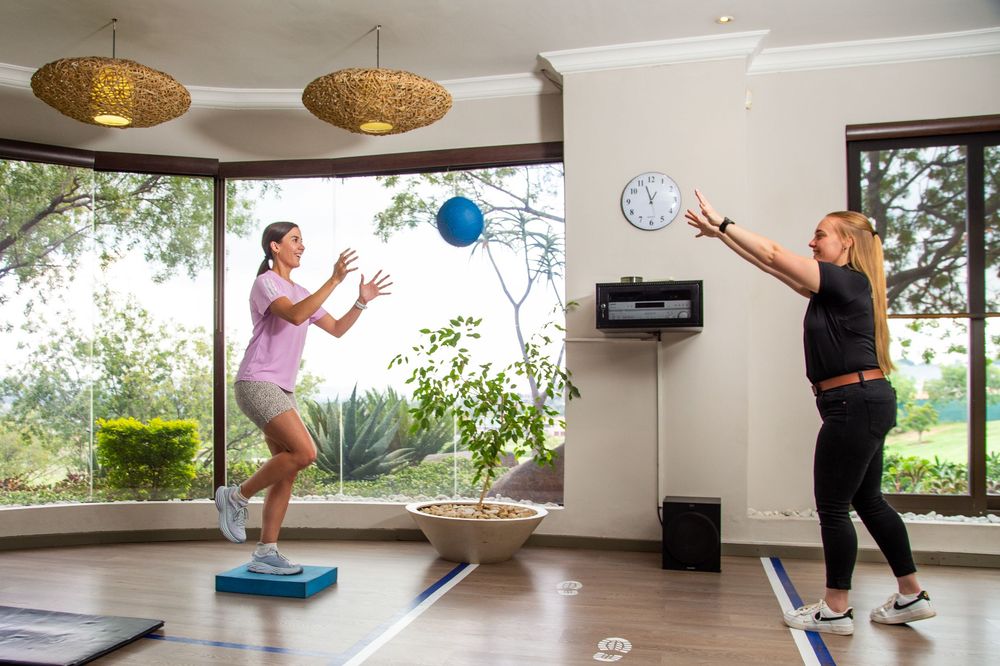 Two women exercising with blue fitness balls on balance boards in a bright indoor workout space with windows.