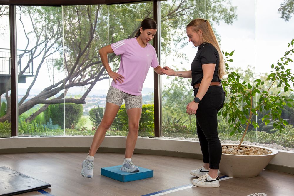 Two people working out together with one balancing on a blue stability training pad while the other supervises the form.