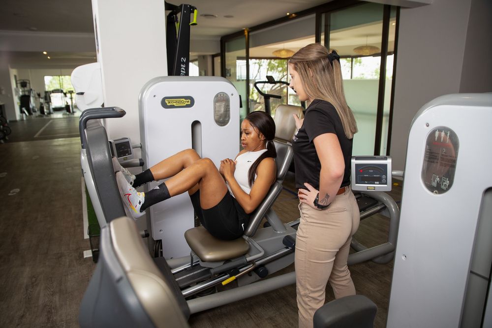 A trainer assists a person using a leg press machine at a modern fitness facility.