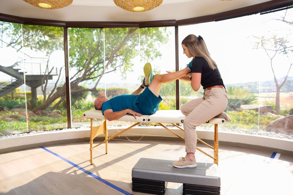 A physical therapist assists a patient performing stretching exercises on a massage table near large windows.